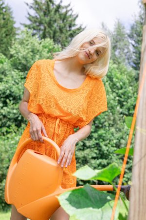 Aphilia bends nude in the garden, orange cloth slipping as she waters plants, skin gleaming under sun — Aphilia #3