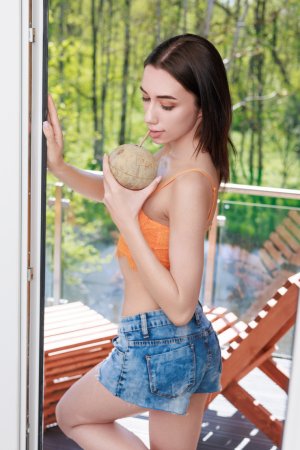 Yeva teases in denim shorts and orange lace, kneeling by window with pineapple and coconut — Yeva #1