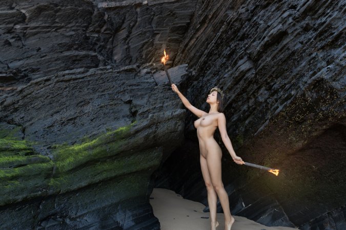 Olivia Linz kneels bare in a cave holding torches, her body lit by fire as she poses solo with crown — Olivia Linz #1