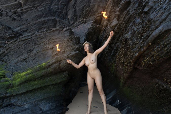 Olivia Linz kneels bare in a cave holding torches, her body lit by fire as she poses solo with crown — Olivia Linz #2