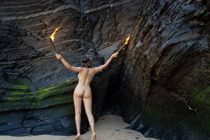 Olivia Linz kneels bare in a cave holding torches, her body lit by fire as she poses solo with crown — Olivia Linz #16